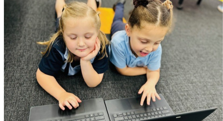 Two students laying on the carpet in the classroom, learning through the use of laptops.