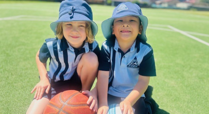 Two students posing for a photo on the field, kneeling down holding a basketball and football.